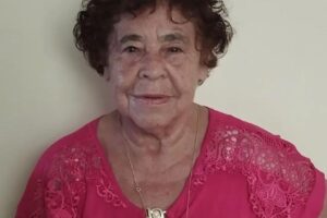 a foto mostra uma ideosa de 70 anos com blusa rosa, cabelos encaracolados e semblante levemente sorrindo