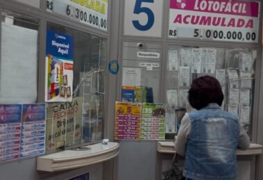 a imagem mostra uma mulher em um a lotérica. De fundo, a banca com anúncios divulgando os prêmios.